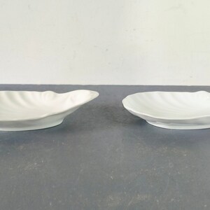 May include: Two white ceramic bowls shaped like seashells. The bowls are smooth and have a glossy finish.
