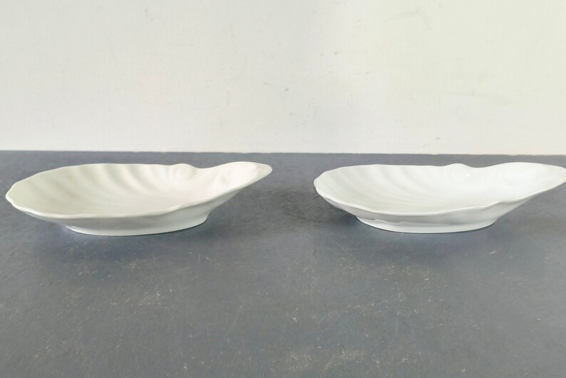 May include: Two white ceramic bowls shaped like seashells. The bowls are smooth and have a glossy finish.