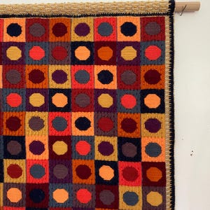 Op de afbeelding: Een kleurrijke, geometrische wandhanger met een herhalend patroon van vierkanten en cirkels. De vierkanten zijn in tinten rood, oranje, geel en blauw, met contrasterende donkere cirkels in het midden. De bovenrand heeft een geweven rand en een houten roede om op te hangen.