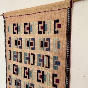Puede incluir: Un tapiz rectangular tejido de tela color canela con textura, suspendido de una varilla de madera. La superficie presenta un patrón de cuadrícula repetitivo de pequeñas formas geométricas en tonos azul marino, azul claro y morado oscuro.