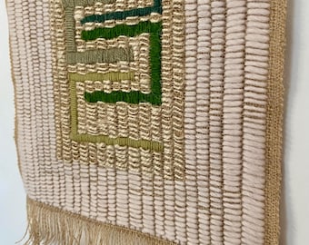May include: A woven wall hanging with a geometric design in shades of green and beige. The piece features a textured weave with a fringe at the bottom. The design is a series of interlocking shapes.
