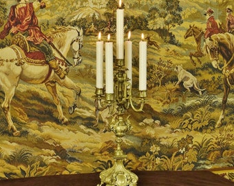 May include: A five-arm brass candelabra with an ornate, detailed base and stem, holding five white taper candles. The candles are lit, with visible flames at the top of each wick, casting a warm glow over the metallic surface of the candle holder.