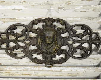 Può includere: Decoro da parete in ghisa ornato con un design decorativo che presenta un medaglione centrale con un viso. Il pezzo è dipinto di colore marrone scuro.