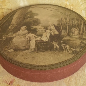 May include: A round, flat box with a deep red base and a lid featuring a sepia-toned illustration of a family scene in a park. The lid is bordered by a dark, textured trim, and the artwork depicts figures in period clothing near trees.