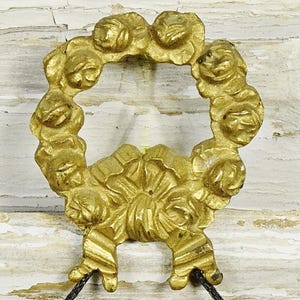 May include: A gold-coloured metal ornament shaped like a circular wreath of roses with a bow at the bottom. The piece features a textured, relief-style floral design throughout the entire circular frame and the central bow detail at the base.