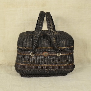May include: A dark brown woven basket with a rectangular shape and a braided base. It features two thick, braided handles attached with silver-toned metal rings and a small central metal clasp. Thin, horizontal yellow accent lines run across the body.