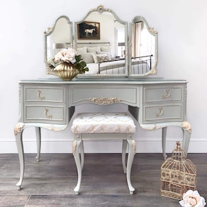 May include: A pale blue vanity table with a tri-fold mirror, drawers, and a matching stool. The vanity has gold detailing. A gold vase with flowers sits on the vanity, and a decorative birdcage is on the floor.