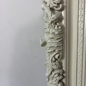Pu&ograve; includere: Primo piano di una cornice per foto bianca ornata con sculture intricate. La cornice &egrave; in legno e ha una finitura anticata.
