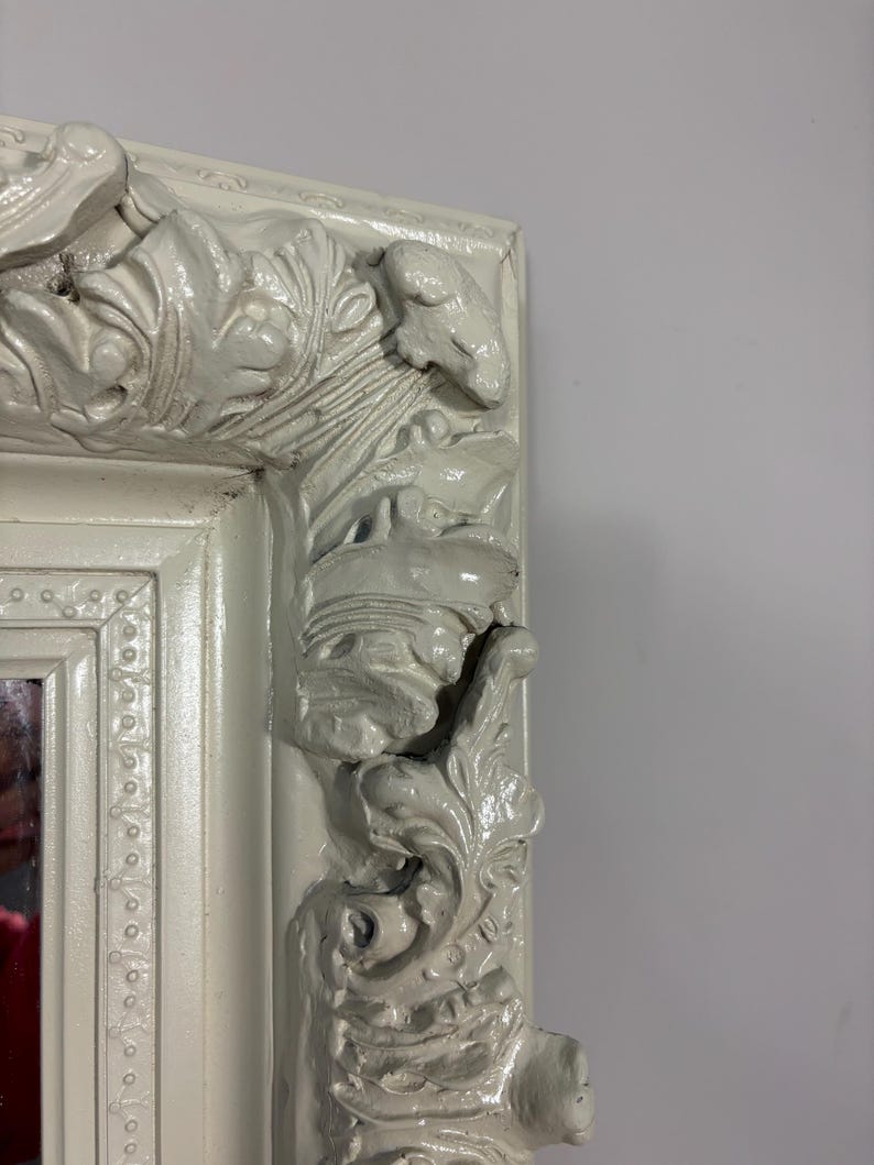 Pu&ograve; includere: Una cornice per specchio bianca ornata con intagli intricati e una finitura usurata.