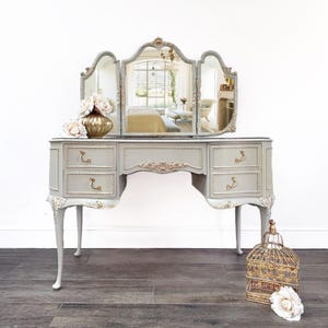 May include: A light grey dressing table with a tri-fold mirror, gold detailing, and decorative drawer handles. The vanity has three drawers on each side and gold trim. A gold vase with white flowers and a decorative birdcage are also present.