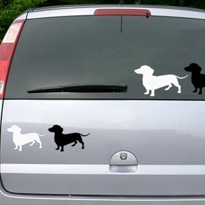 May include: Car window decals featuring dachshund dog silhouettes in white and black. The decals are displayed on a car's rear window, with three dogs at the top and two at the bottom. The background is a blurred view of green foliage.