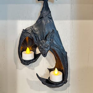May include: A black wall-mounted bat sculpture featuring two integrated circular holders for tea light candles. The bat is depicted hanging upside down with its wings spread, and each wing tip contains a small white battery-operated candle.