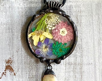 May include: A round pendant necklace featuring a clear resin dome filled with pressed flowers. The flowers are yellow, pink, purple, and green. The pendant is attached to a black leather cord and has a small, oval-shaped, pale yellow stone hanging from the bottom.