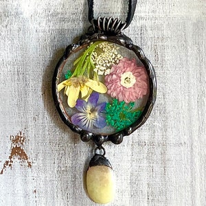 May include: A round pendant necklace with a clear resin dome containing pressed flowers. The flowers are yellow, pink, purple, and green. The pendant is attached to a black leather cord and has a small, oval-shaped, light yellow stone hanging from the bottom.