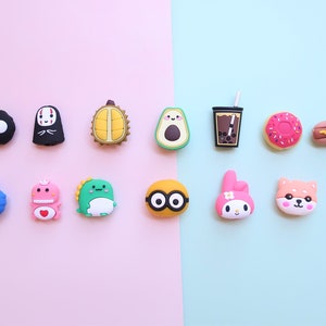 May include: A collection of 14 colorful silicone phone charms featuring various characters and objects, including a black No Face character, a green dinosaur, a blue Cookie Monster, a pink Hello Kitty, a brown Shiba Inu, a rainbow, a durian, an avocado, a boba tea, a donut, and a hot dog.