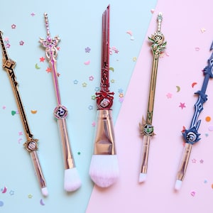 May include: Five makeup brushes with sword-shaped handles. The handles are decorated with gold, silver, and red accents. The brushes are arranged on a pink and blue background with confetti.