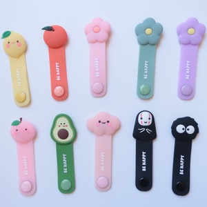 May include: Ten colorful cable organizers with different cartoon characters. Each organizer has the text "BE HAPPY" printed on it.