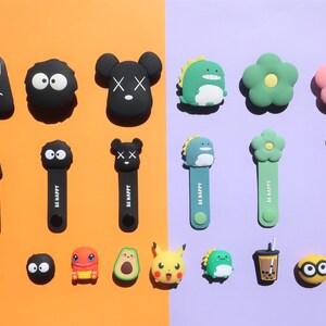 May include: Assortment of colorful cartoon character cable organizers.  The characters include No Face, Totoro, Mickey Mouse, a dinosaur, a flower, a Pikachu, an avocado, a frog, a boba tea cup, and a Coca-Cola can.  The organizers are made of silicone and have the words "Be Happy" printed on them.