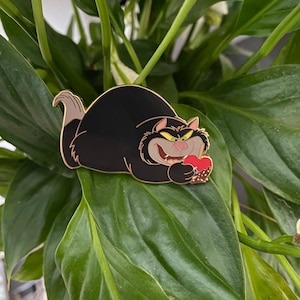 Op de afbeelding: Een zwarte kat met groene ogen en een ondeugende grijns houdt een rode appel vast met witte spikkels. De kat ligt op een groen blad.