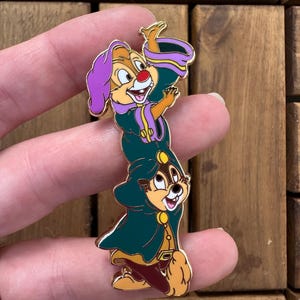 Op de afbeelding: Emaille pin met twee cartoon chipmunks in groene en paarse outfits. De bovenste chipmunk houdt een gouden hoorn vast, de onderste glimlacht. De pin heeft een gouden rand. De achtergrond is van hout.