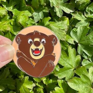 Peut inclure: Un personnage de dessin animé d'ours brun avec un grand sourire et les bras croisés, sur un fond de couleur pêche. L'ours porte un manteau de fourrure brun et a un museau blanc.