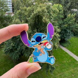 Op de afbeelding: Blauwe Stitch-figuur emaille pin met een paars oor, die een peperkoekman, een kerstboom en een kerstman vasthoudt.