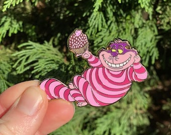 May include: Pink and white striped Cheshire Cat pin with a purple cupcake in its paw. The pin is made of metal and has a shiny finish.