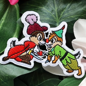May include: An embroidered patch featuring two cartoon chipmunks engaged in a sword fight. One chipmunk wears a red cape and hat with a pink feather, while the other wears a green tunic and hat. Both figures are outlined in white thread.