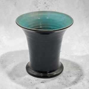 May include: A ceramic vase with a flared rim and a tapered body. The exterior features a smooth, dark charcoal-black glaze, while the interior is finished in a contrasting vibrant turquoise-blue glaze. The vessel has a wide, stable circular base.