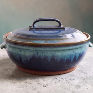 Op de afbeelding: Een ronde keramische ovenschaal met bijpassend deksel, voorzien van een glanzend glazuur dat verloopt van diep marineblauw aan de basis naar een lichtere turquoise en blauwe kleur op de bovenrand en het deksel, met een natuurlijke terracotta voet.