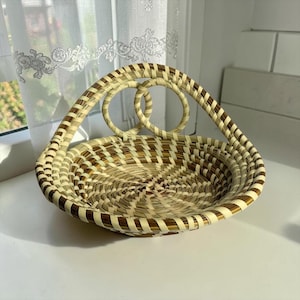 May include: A round woven basket featuring a natural tan and dark brown striped pattern. The basket has a shallow, circular base and a unique handle design consisting of two interconnected circular loops that rise from the rim of the basket.