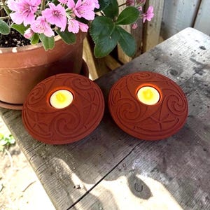 May include: Two round, terracotta-colored candle holders featuring intricate, embossed swirl and floral patterns. Each holder has a central circular opening containing a small, lit white tea light candle with a warm yellow flame.