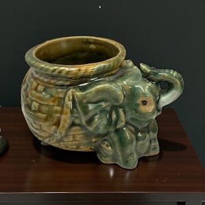 Peut inclure: Pot de fleurs en céramique en forme d'éléphant, avec une texture de panier sur le corps et un glaçage vert et marron. La trompe de l'éléphant forme une poignée, et le pot a une large ouverture.