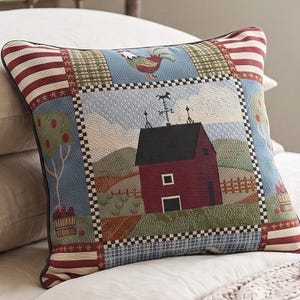 May include: A square throw pillow featuring a woven tapestry design of a red barn with a black roof and a weather vane. The scene includes a rooster, a fruit tree, and a basket of apples, framed by a checkered border and red and white stripes.
