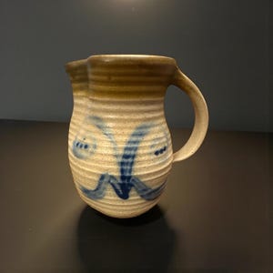 May include: A handmade ceramic pitcher with a tan and blue color scheme. The pitcher has a ribbed texture and a curved handle. Blue abstract designs decorate the body of the pitcher. The pitcher is suitable for serving beverages.