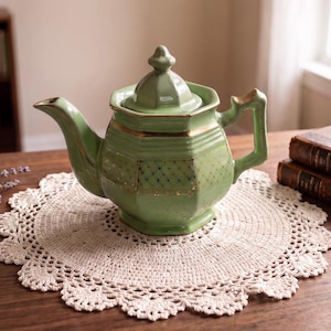 May include: A sage green ceramic teapot with an octagonal body, a matching lid, and a gold-colored starburst pattern around the center. The teapot features a curved spout and a handle, with gold trim accents along the rim, lid, and base edges.