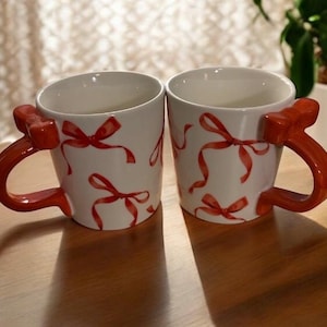May include: Two white ceramic mugs with red bow handles and red bow accents. The mugs have a repeating pattern of red bows. The mugs are on a wooden surface.