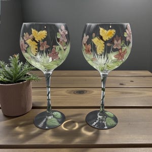 May include: Two clear wine glasses with a painted butterfly and floral design. The butterflies are yellow, and the flowers are pink and red. The stems are green with leaf accents, and the bases are round and gray.