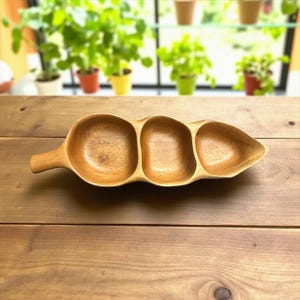 May include: A wooden serving dish with three connected compartments and a handle. The dish is made of light brown wood and has a smooth finish. The compartments are oval-shaped and appear to be empty. The dish is on a wooden table.