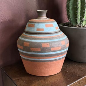 May include: A terracotta-colored ceramic jar with a lid. The jar features a geometric pattern of blue and orange rectangles and stripes on a dark brown background. The jar is displayed on a wooden surface.