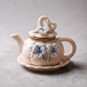May include: A cream-coloured porcelain teapot-shaped trinket box featuring hand-painted blue pansy flowers on the body and lid. The lid is hinged with a gold-tone metal band and a bow-shaped clasp, resting on a matching scalloped saucer base.