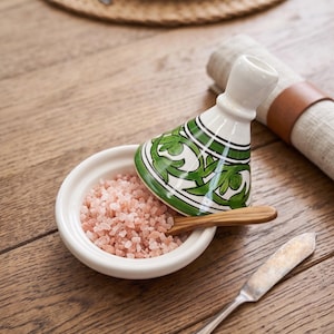 May include: A ceramic tagine-shaped salt cellar with a white base and a conical lid featuring green hand-painted floral patterns. The bowl is filled with coarse pink Himalayan salt and includes a small wooden spoon resting inside the container.