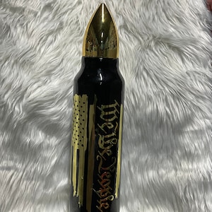 May include: Black tumbler with a gold-colored bullet-shaped lid. The tumbler features a gold-colored American flag design and the words "We the People" in gold script. The tumbler is cylindrical and appears to be made of metal.