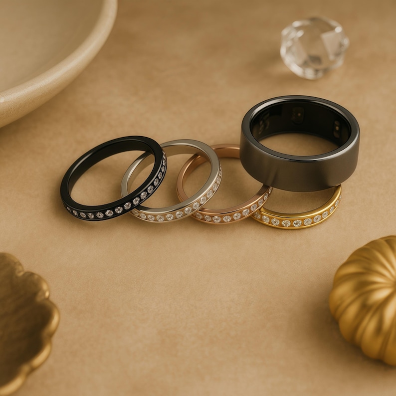 Puede incluir: Una colecci&oacute;n de anillos en varios colores, incluyendo negro, plateado, oro rosa y dorado. Algunos anillos est&aacute;n adornados con peque&ntilde;as piedras preciosas transparentes. Un anillo grande de color gris oscuro se encuentra encima de los dem&aacute;s. Los anillos se exhiben sobre una superficie beige.