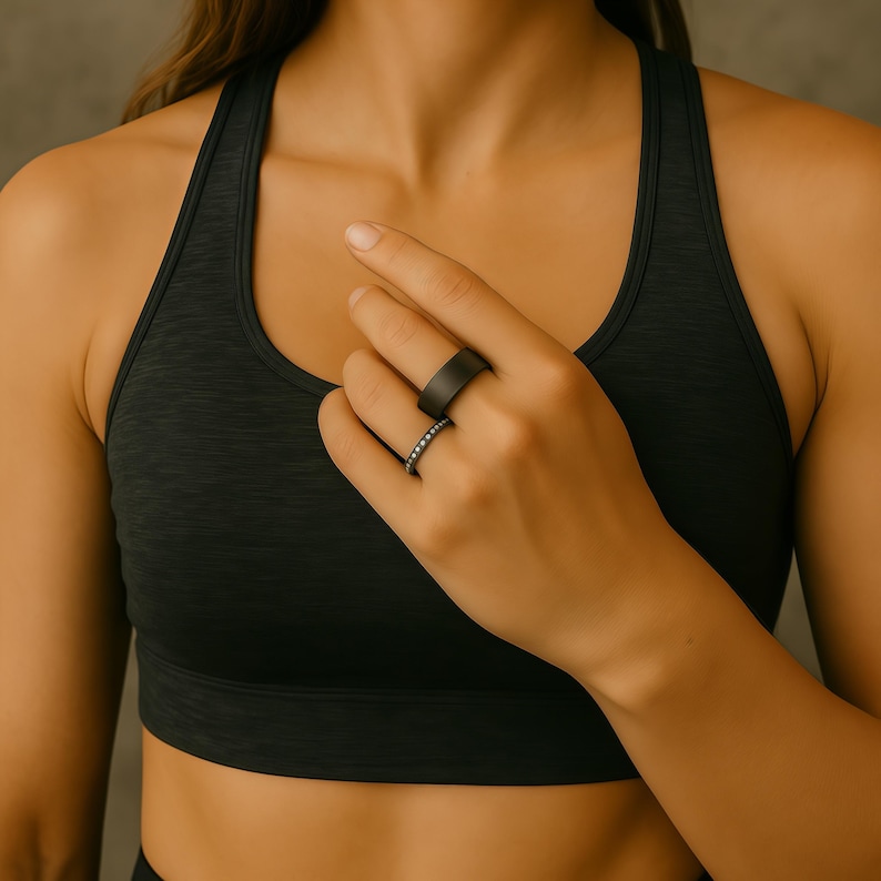 Puede incluir: Primer plano de dos anillos negros. Uno es una banda ancha, el otro una banda m&aacute;s delgada con peque&ntilde;as piedras brillantes. Los anillos est&aacute;n en una mano, con un sujetador deportivo gris oscuro en el fondo.