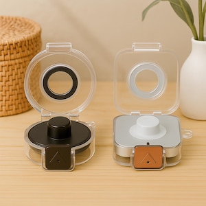 May include: Two clear, hinged cases. One has a circular design with a black interior and button, the other a square design with a silver interior and white button. Both have a small, brown square with a triangle.