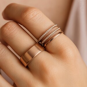 Puede incluir: Una mano muestra una colecci&oacute;n de anillos de oro. Los anillos incluyen una banda ancha, una banda delgada y dos bandas adornadas con peque&ntilde;as piedras brillantes. Los anillos son de color oro c&aacute;lido.