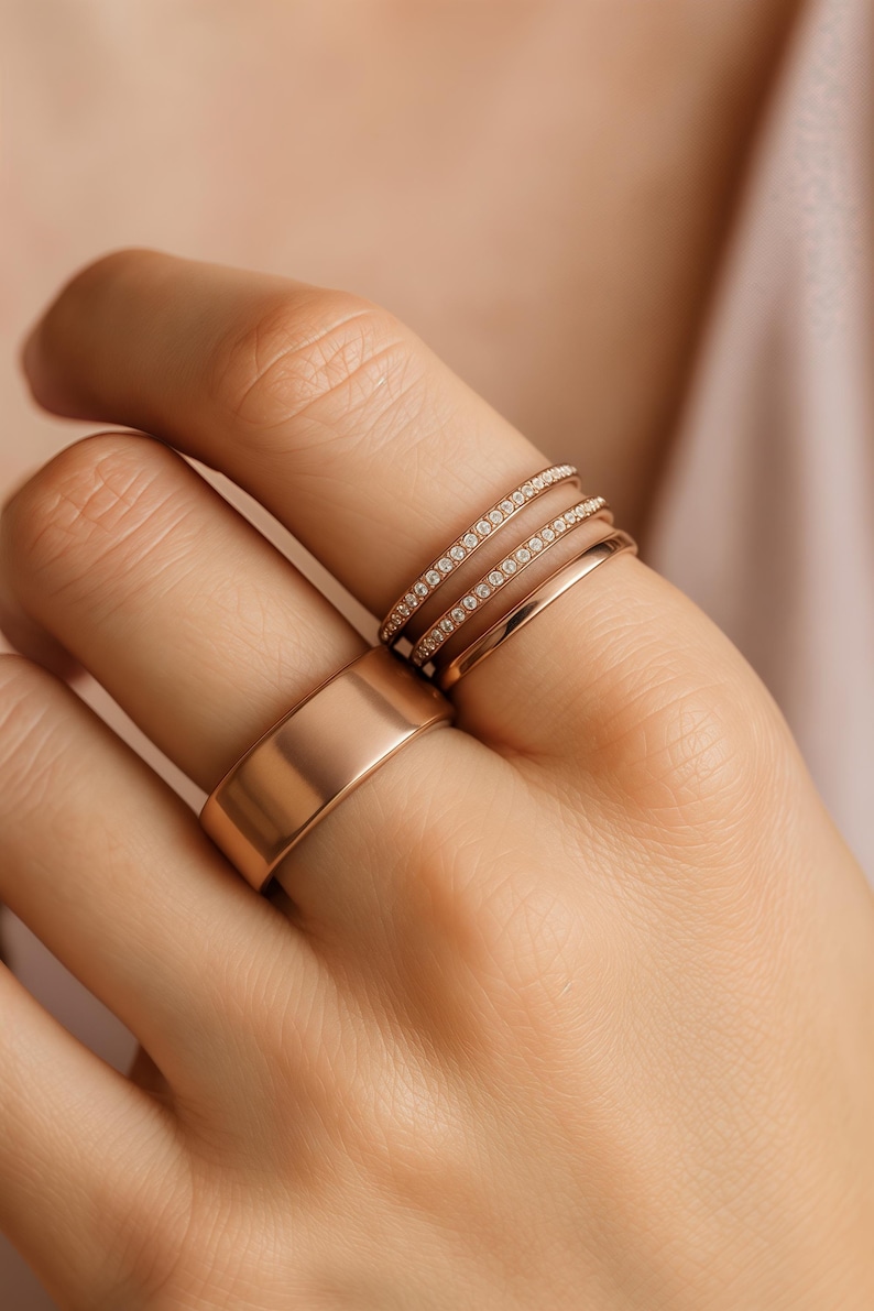 Puede incluir: Una mano muestra una colecci&oacute;n de anillos de oro. Los anillos incluyen una banda ancha, una banda delgada y dos bandas adornadas con peque&ntilde;as piedras brillantes. Los anillos son de color oro c&aacute;lido.