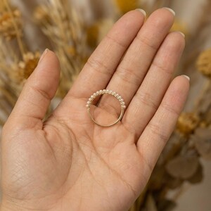 May include: A delicate gold ring featuring a band of small, round pearls. The ring is held in a hand, showcasing its elegant design and craftsmanship. The pearls are evenly spaced, creating a classic and timeless look.