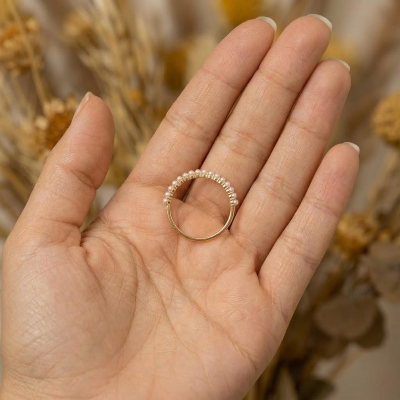 May include: A delicate gold ring featuring a band of small, round pearls. The ring is held in a hand, showcasing its elegant design and craftsmanship. The pearls are evenly spaced, creating a classic and timeless look.